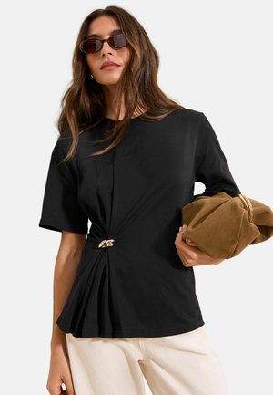 Nő sötét napszemüveget visel, fekete rövid ujjú felsőt arany csattal, világosbarna puha táskát tart, bézs nadrágot visel, semleges háttér előtt.