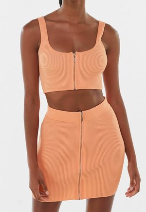 Femme portant une tenue deux pièces côtelée de couleur pêche avec des fermetures éclair à l'avant sur le crop top et la mini-jupe, debout devant un fond uni.