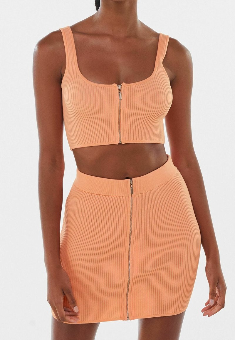 Femme portant une tenue deux pièces côtelée de couleur pêche avec des fermetures éclair à l'avant sur le crop top et la mini-jupe, debout devant un fond uni.