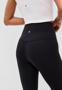 Nahaufnahme einer Person, die schwarze Leggings mit hohem Bund und ein weißes kurzes Oberteil trägt, beide mit einem kleinen reflektierenden Logo auf der Rückseite.