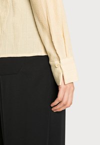 Camisa bege texturada com tecido canelado, punhos abotoados e um corte descontraído. Combinada com calças pretas, exibindo um estilo limpo e minimalista.