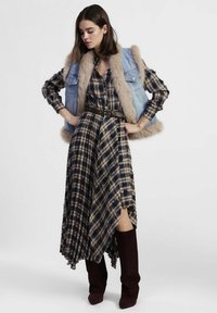 Gilet di denim con rifinitura in pelliccia sopra un abito midi a quadri. L'abito presenta un orlo asimmetrico e una cintura in vita. Stivali bordeaux al ginocchio completano il look.