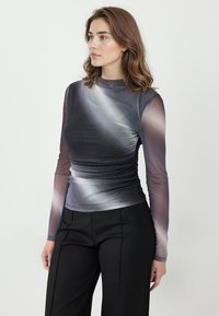 Młoda kobieta o falowanych brązowych włosach, ubrana w dopasowany, długorękawowy top w szaro-białym gradientcie oraz czarne spodnie, stojąca na jednolitym tle.