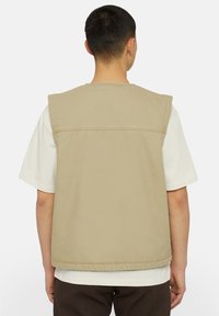 Beige ermeløs vest med et enkelt design, preget av en rett nederkant og en sømløs bakside, lagd over en hvit T-skjorte.