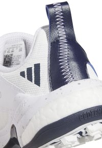 Bílá sportovní bota s námořnicky modrým jazýčkem na patě, texturovaným svrškem z látky a viditelným prošíváním. Má polstrovanou podrážku s logem.