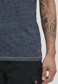 Granatowy top bez rękawów z teksturowanym wykończeniem i brązową naszywką z logo. Zbliżenie na dłoń z tatuażem na ramieniu, noszącą czarne spodnie.