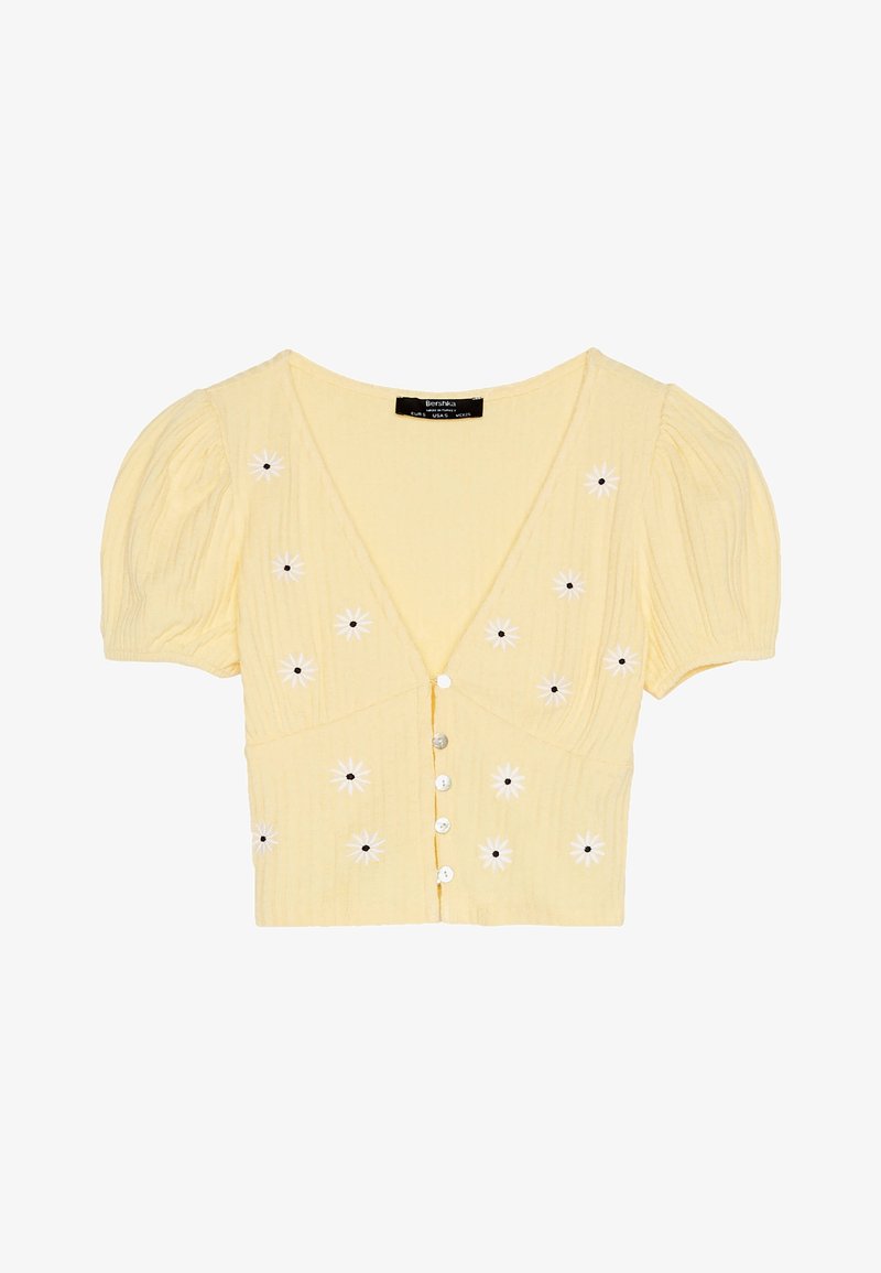 Cardigan court jaune avec manches ballon, col en V et cinq boutons à l'avant. Présente une broderie de fleurs blanches avec des centres noirs sur le devant.