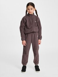 Dziewczyna z warkoczami, ubrana w dopasowaną ciemnobrązową polarową kurtkę na zamek i spodnie, stoi z rękami swobodnie opuszczonymi wzdłuż ciała.