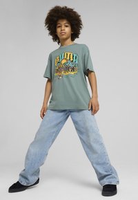 Camisa de manga corta verde con un diseño gráfico colorido, combinada con jeans de pierna ancha de color azul claro y zapatos negros.