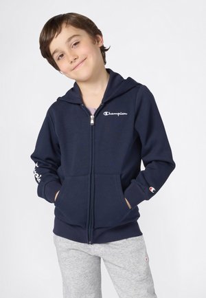 Jeune garçon souriant, portant un sweat à capuche zippé bleu marine avec le logo "Champion" et un pantalon de survêtement gris clair, mains dans les poches.