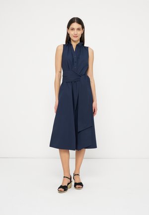 Naine seisab valgel taustal, kandes tumesinist varrukateta midi-mantlit, millel on nööpidega krae, ning musti kiiljalanõusid.