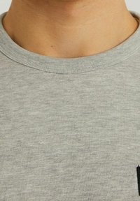 Maglietta grigia in cotone con scollo rotondo e una texture sottile. Presenta una piccola tasca nera sul lato sinistro.