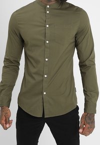 Camicetta verde oliva a maniche lunghe con colletto chino, tasca frontale e bottoni bianchi; texture liscia e design aderente.