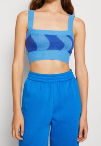 Pletený modrý top s krátkým střihem a čtvercovým výstřihem, širokými ramínky a abstraktními vzory v tmavší modré barvě. V páru s odpovídajícími modrými tepláky.