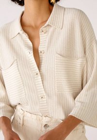 Krémová proužkovaná košile s předním zapínáním na knoflíky, dvěma náprsními kapsami a širokými rukávy. Tkanina vypadá texturovaně a lehce.