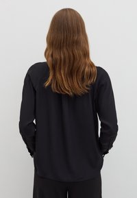 Black long-sleeve blouse made of smooth fabric, featuring buttoned cuffs and a curved hem, viewed from the back.