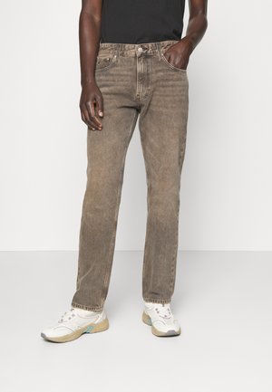 Person trägt verblasste braune Jeans mit geradem Bein, schwarzes Oberteil, weiße Turnschuhe und steht mit einer Hand in der Tasche vor einem einfarbigen Hintergrund.