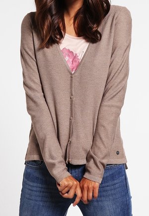 Kvinde iført en knaplukket taupe cardigan over en lyserød blomstret skjorte og blå jeans, stående med hænderne foldet foran.
