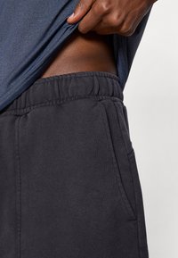 Calças de fato pretas com um cós elástico, bolsos laterais e uma textura suave, parcialmente cobertas por uma camisa azul.