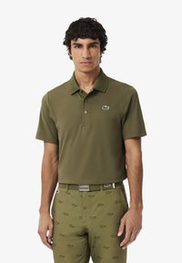 Vihreä oliivinvärinen polo-paita, jossa on klassinen kaulus ja lyhyet hihat. Rinnassa on pieni vihreä Lacoste-logo brodeerattuna.