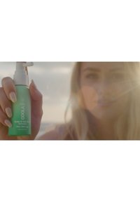 Zielona butelka ze sprayem oznaczona napisem "Mgiełka do skóry głowy i włosów" z białą pompką, trzymana w dłoni z długimi, jasnymi paznokciami. Tło lekko rozmyte.