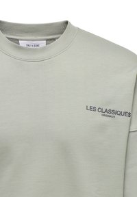 Hellgrüner Kapuzenpullover mit geripptem Rundhalsausschnitt, weichem Material und dem gedruckten Text "LES CLASSIQUES ORIGINAUX" in Dunkelblau auf der Brust.