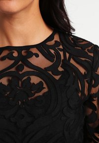 Sort gennemsigtig top med detaljeret sort blomsterbroderi, rund halsudskæring og teksturerede detaljer. Elegant og tætsiddende design.