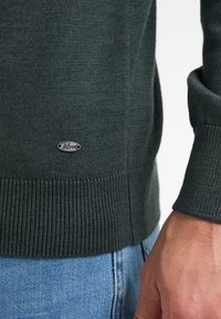 Pulover tricotat de culoare verde închis, cu manșete și tiv cu nervuri, având un mic etichetă logo argintie pe lateral. Textură moale și design ajustat.