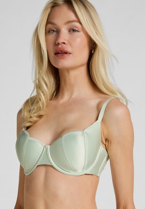 Blonde Frau, die einen hellgrünen Satin-BH mit Bügeln trägt, schaut mit neutralem Ausdruck nach vorne vor einem schlichten grauen Hintergrund.