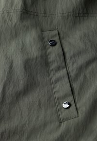 Oliivivihreä kangas, jossa on hieman ryppyinen rakenne, ja siinä on pystysuora läpän tasku, joka on kiinnitetty kahdella hopeanvärisellä nepparilla.