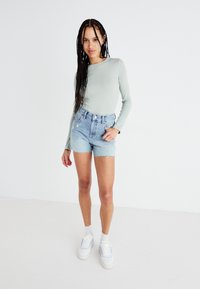 Haut à manches longues vert clair associé à un short en denim taille haute avec un ourlet effiloché et des détails floraux brodés, porté avec des baskets blanches.