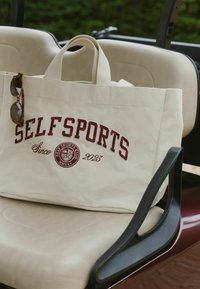 Borsa tote in tela beige con testo ricamato color amaranto "SELF SPORTS" e logo. Dotata di manici superiori robusti. Occhiali da sole appoggiati sul bordo della borsa.
