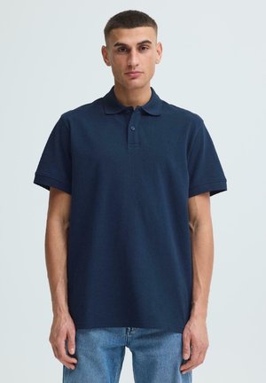 Young man with short hair wearing a navy blue polo shirt and light blue jeans, standing against a plain light background.