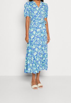 Kobieta ubrana w niebieską, kwiatową sukienkę midi z guzikami z przodu, bufkami na rękawach i paskiem w talii, w połączeniu z kremowymi sandałami na klocku z odkrytymi palcami.