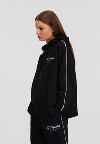Zwarte hoodie met trekkoord, zijden witte strepen en logo-opdruk aan de voorkant. Zachte stofstructuur, comfortabele pasvorm, gecombineerd met bijpassende broek.