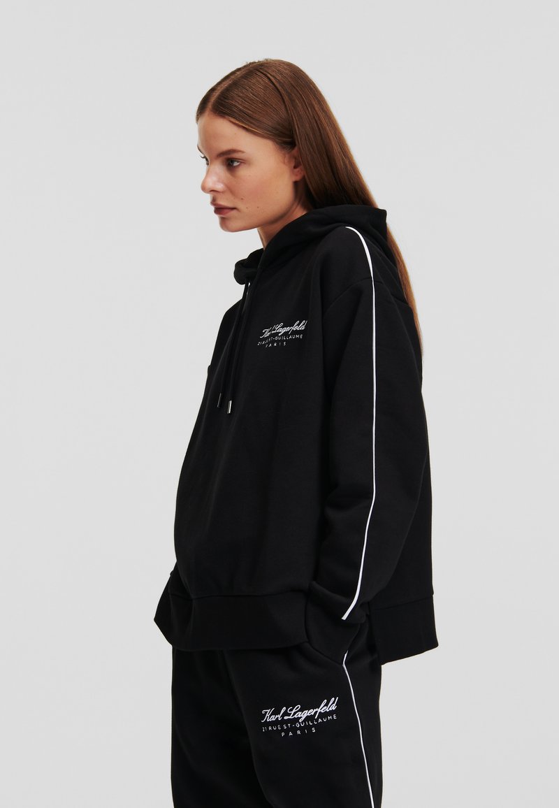 Schwarzer Hoodie mit Kordeln, seitlichen weißen Streifen und Logo-Details auf der Vorderseite. Weicher Stoff mit entspannter Passform, kombiniert mit passenden Hosen.