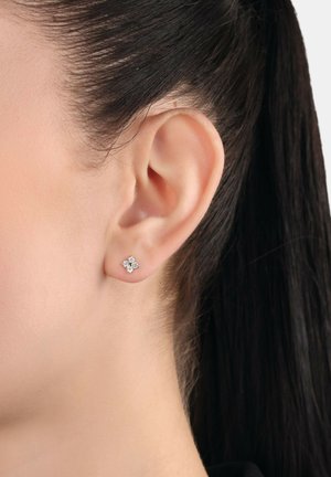 Primo piano dell'orecchio di una donna con capelli scuri, che indossa un piccolo orecchino a perno d'argento a forma di fiore a quattro petali con pietre trasparenti.