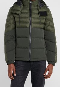 Mand iført en olivengrøn farveblokeret polstret jakke med lynlås fortil og sorte jeans foran en grå baggrund.