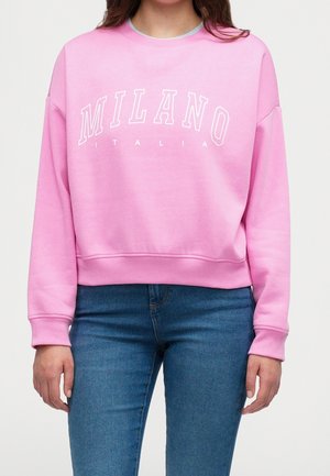 Kvinde iført en knaldrød lyserød sweatshirt med teksten "MILANO ITALIA" og blå jeans, vist fra skuldre til hofter mod en ensfarvet baggrund.