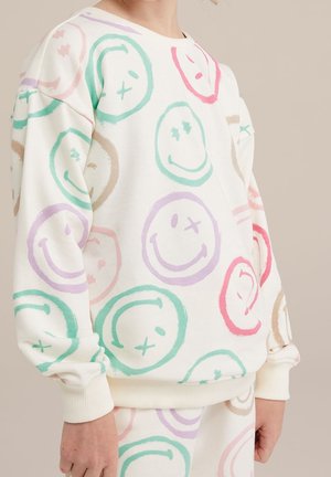 Enfant portant un sweat-shirt blanc et un pantalon avec des motifs de visages souriants clignant de l'œil multicolores en tons de vert, violet, rouge et beige.