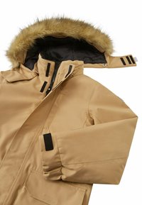 Giacca isolante beige con cappuccio foderato in pelliccia, chiusure con zip e bottoni a scatto, numerose tasche e dettagli in Velcro nero sulle maniche.