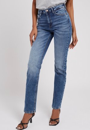 Kvinde iført højtaljede, to-tone blå straight-leg jeans kombineret med sorte sandaler med hæl og en lysegrå top, stående mod en ensfarvet baggrund.