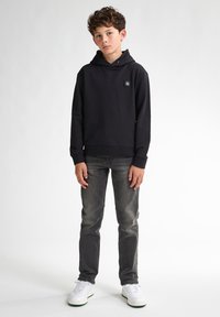 Felpa nera con un patch logo sul davanti, abbinata a jeans grigi e sneaker bianche. La felpa ha una vestibilità ampia e una tasca a marsupio.
