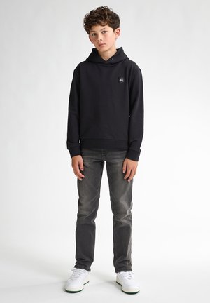 Zwarte hoodie met een logo patch aan de voorkant, gecombineerd met grijze jeans en witte sneakers. De hoodie heeft een relaxed fit en een kangaroo pocket.