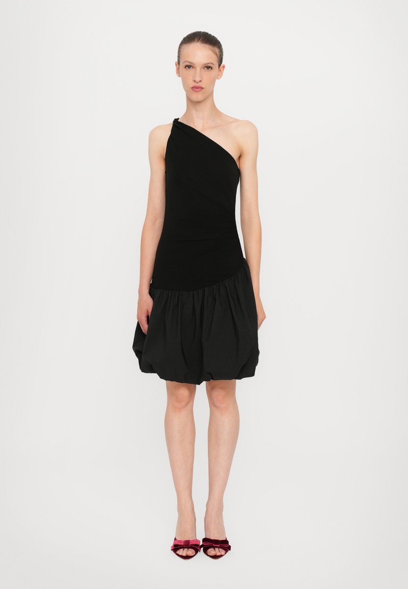 Robe noire à une épaule avec un corsage ajusté, un tissu texturé et une jupe volumineuse. Associée à des chaussures à talons en velours rouge.