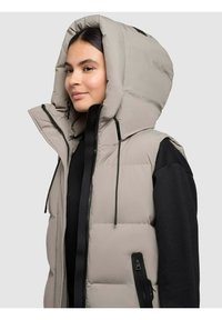 Beež puffer vest, millel on suur kapuuts. Omab lukuga esitaskut, küljetaskuid ning kontrastseid musti detaile. Sujuv, isoleeritud tekstuur.