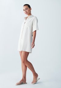 Svetlo bež oblečna košulja s kratkimi rokavi, narejena iz mehke tkanine, z gumbnim zapiranjem in žepom na prsih, z udobnim krojem in stranskimi razporki.