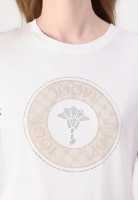 Bílé bavlněné tričko s kulatým designem a béžovými a stříbrnými ozdobami, včetně květinového motivu ve středu.