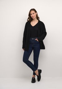 Schwarze, flauschige Strickjacke mit offener Vorderseite, über einem schwarzen Oberteil und dunkelblauen Skinny-Jeans getragen. Das Modell steht mit einem Bein überkreuzt in schwarzen Loafers.