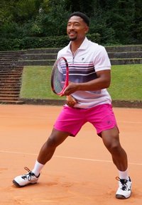 Tennis player in a striped, white polo and bright pink shorts, holding a red tennis racket, poised on a clay court.